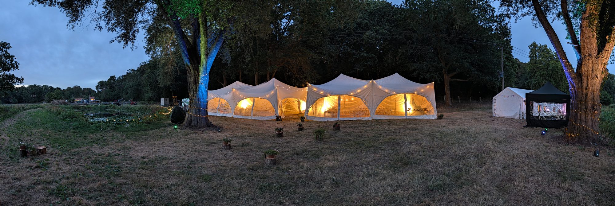 Marquees glowing with festoon lights at dusk in a field edged with trees